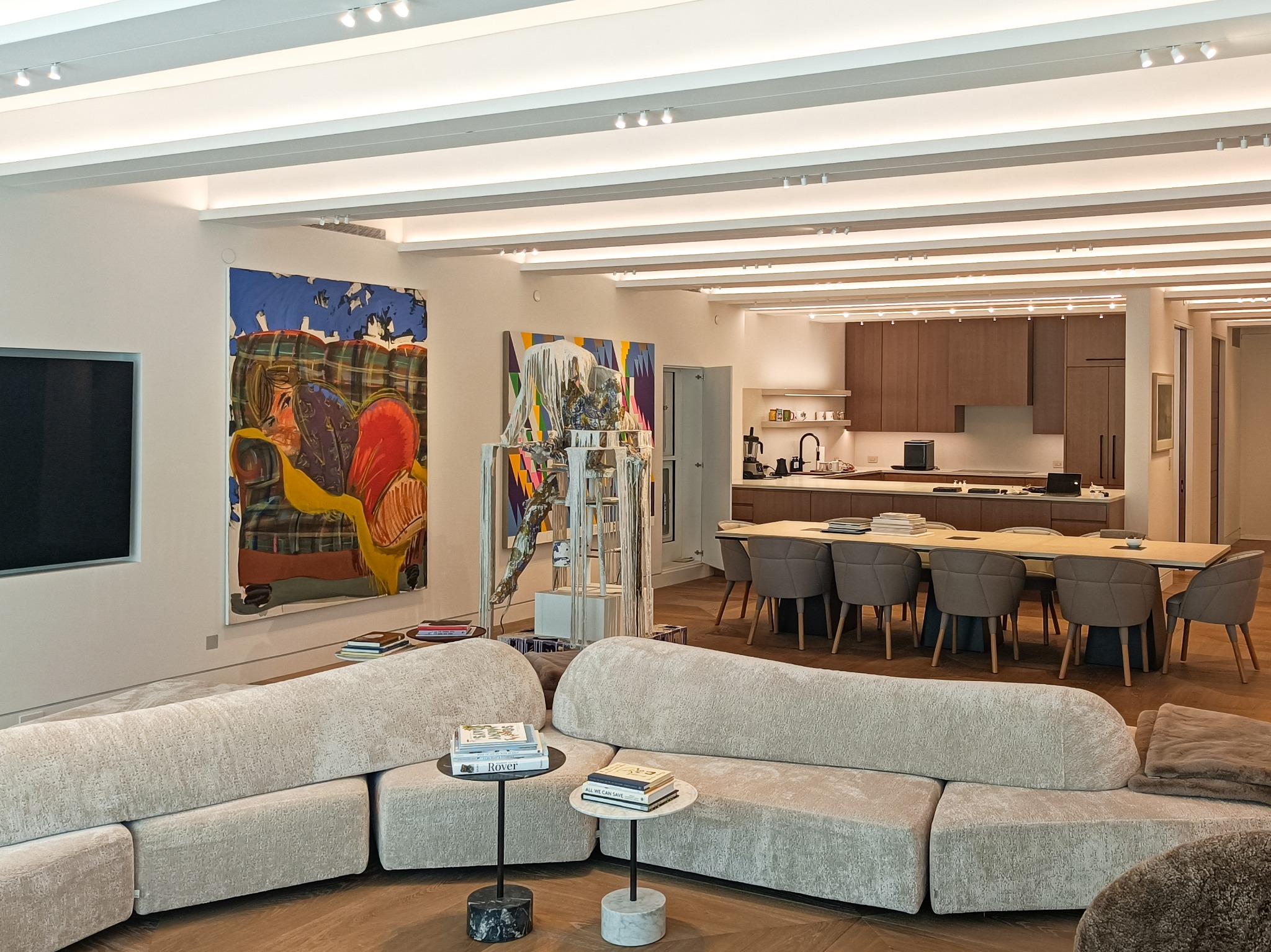 Nowoczesne wnętrze salonu z oświetleniem sufitowym typu spotlight, jasną sofą modułową, stolikami pomocniczymi i widoczną kuchnią z jadalnią w tle.