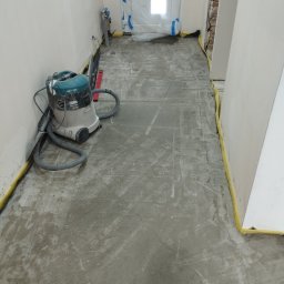 Mateusz Kuleta - Przygotowanie podłoża: Szlifowana wylewka samopoziomująca w korytarzu, zabezpieczone ściany taśmą malarską, odkurzacz przemysłowy Makita. Widoczne ślady po szlifowaniu.