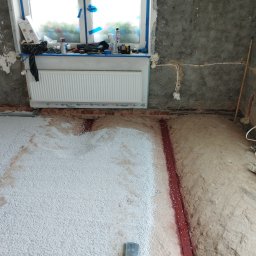 Mateusz Kuleta - Nierówna podłoga w trakcie wylewania izolacji z białego granulatu, widoczne ślady po instalacjach w podłożu, ściany w surowym stanie, okno zabezpieczone folią malarską, grzejnik zamontowany.
