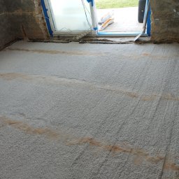 Mateusz Kuleta - Wyrównana, szara, chropowata powierzchnia podłogi z widocznymi smugami zabrudzeń, przygotowana pod dalsze prace wykończeniowe, przy oknie widoczne worki z materiałem i przedłużacz.