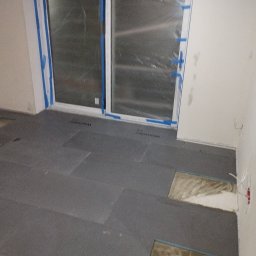 Mateusz Kuleta - Przygotowanie podłoża pod panele: rozłożone płyty izolacyjne na podłodze pomieszczenia z zabezpieczonymi folią i taśmą oknami balkonowymi, widoczne fragmenty starej podłogi i wystające kable...