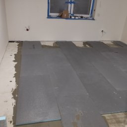 Mateusz Kuleta - Układanie paneli podłogowych w pomieszczeniu, widoczne płyty izolacyjne Warmtec i warstwa kleju na podłodze, okno zabezpieczone taśmą malarską.