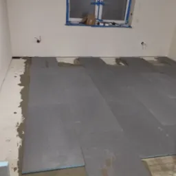 Układanie paneli podłogowych w pomieszczeniu, widoczne płyty izolacyjne Warmtec i warstwa kleju na podłodze, okno zabezpieczone taśmą malarską.