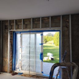 Mateusz Kuleta - Wnętrze w trakcie remontu z widoczną konstrukcją metalową ścian działowych, zabezpieczonymi folią drzwiami balkonowymi z widokiem na ogród, oraz drabina aluminiowa.