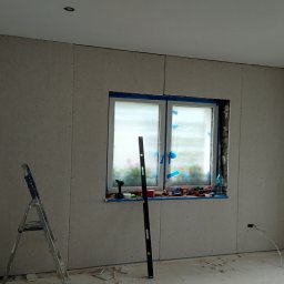 Mateusz Kuleta - Pokój w trakcie prac wykończeniowych: ściany obłożone płytami gipsowo-kartonowymi, okno zabezpieczone taśmą malarską, poziomica oparta o ścianę, drabina rozstawiona w rogu, widoczne narzędzia...