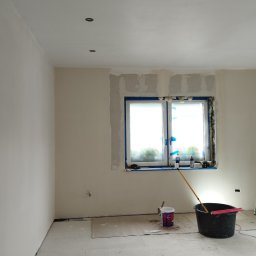 Mateusz Kuleta - Pokój w trakcie remontu: ściany po szpachlowaniu, zabezpieczone okno folią malarską, wiadro z farbą, drabina aluminiowa i widoczne ślady prac remontowych.