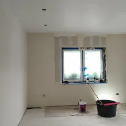 Pok&oacute;j w trakcie remontu: ściany po szpachlowaniu, zabezpieczone okno folią malarską, wiadro z farbą, drabina aluminiowa i widoczne ślady prac remontowych.