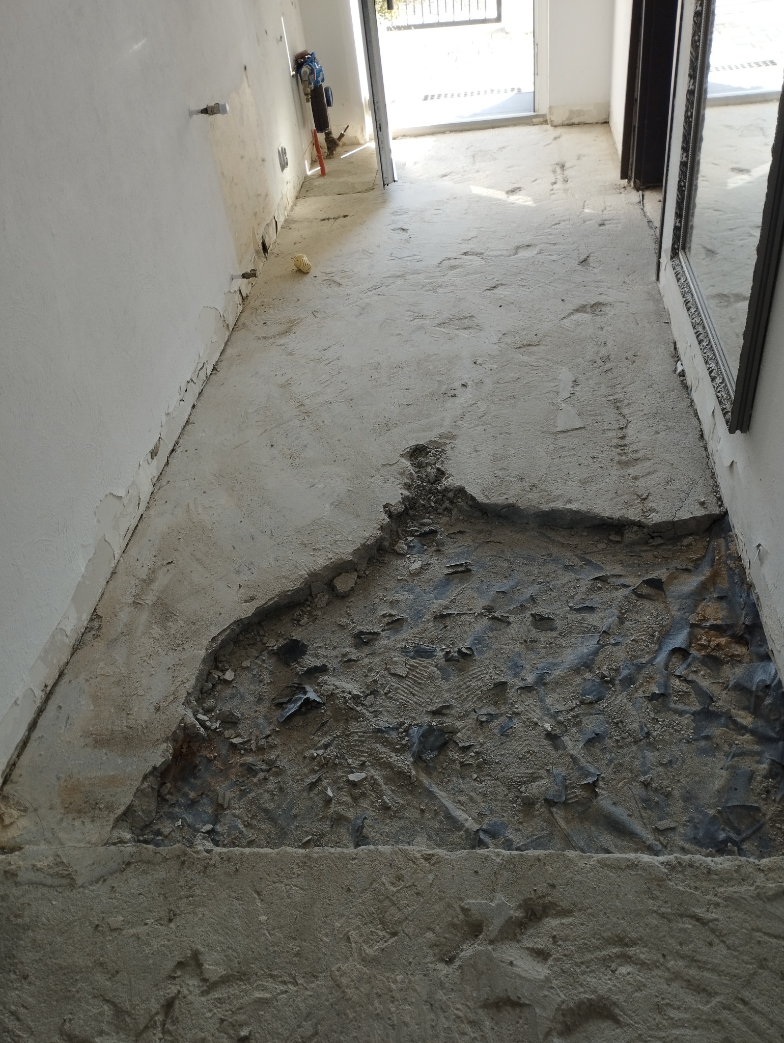 Fragment podłogi w trakcie remontu z usuniętą wylewką, odsłaniający warstwę gruzu i folii, ściany z widocznymi śladami po skuciu, widoczne rury instalacyjne i narzędzia w tle.