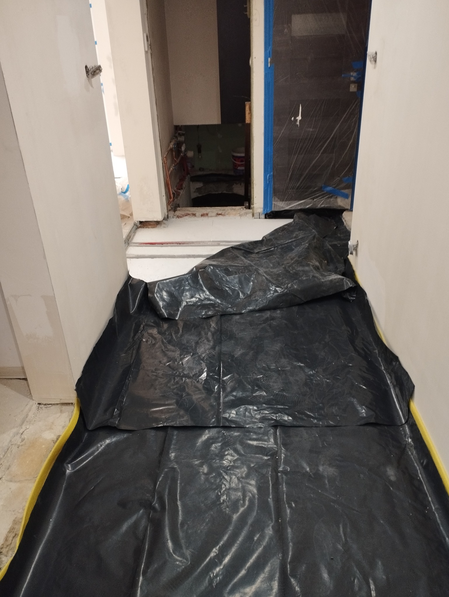 Przygotowanie podłoża do prac wykończeniowych: czarna folia ochronna rozłożona na podłodze z żółtą taśmą zabezpieczającą przy ścianach, widoczne otwarte drzwi do łazienki z zabezpieczonymi folią...