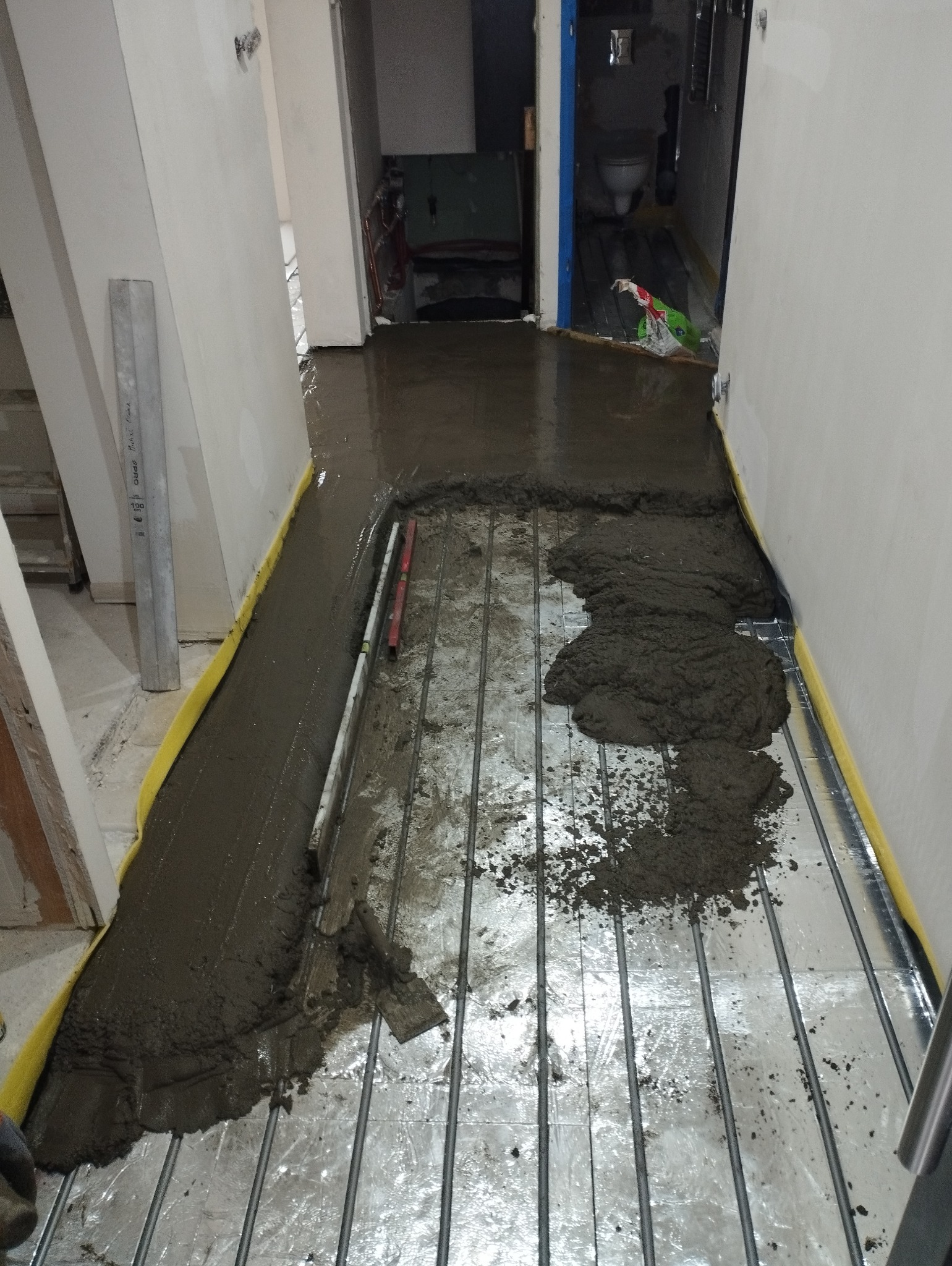 Wylewka samopoziomująca rozprowadzana na folii z ogrzewaniem podłogowym, widoczne narzędzia i poziomica.