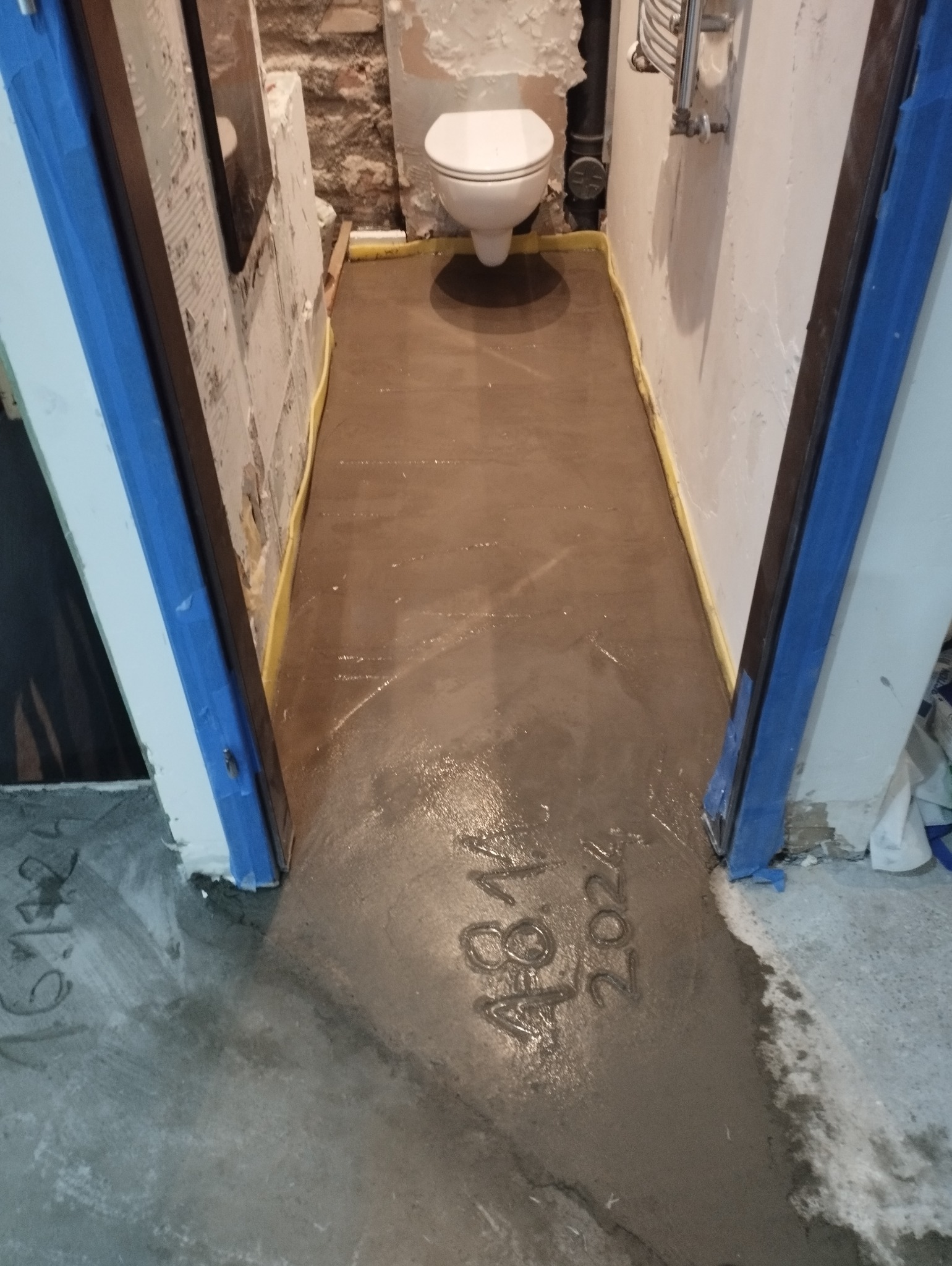 Świeżo wylana, jeszcze mokra wylewka samopoziomująca w remontowanej łazience z toaletą, zabezpieczona żółtą folią przy ścianach i niebieską taśmą malarską przy ościeżnicy, z odciśniętą datą 4.8.14...