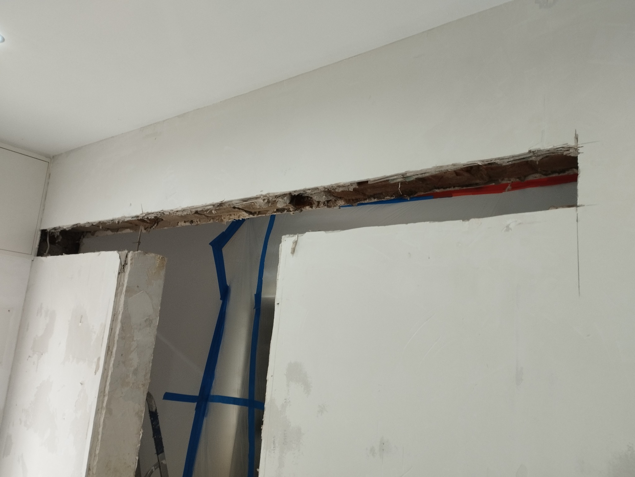 Przygotowanie ściany do prac wykończeniowych: wycięty otwór w ścianie z widocznymi instalacjami zabezpieczonymi niebieską taśmą malarską.