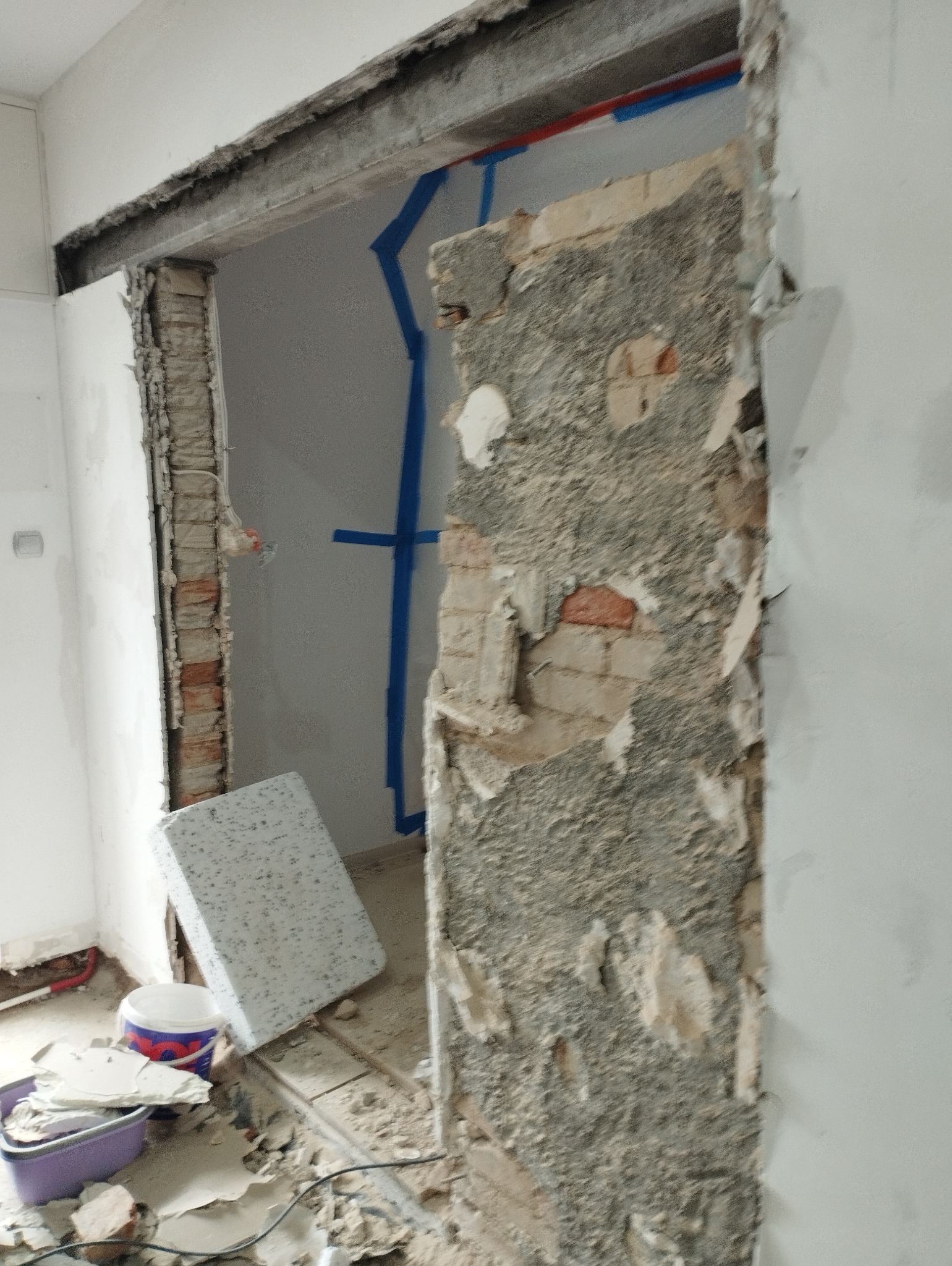 Demontaż ościeżnicy drzwiowej z odsłoniętą cegłą i fragmentami starej zaprawy, widoczne ślady taśmy malarskiej, gruz i narzędzia na podłodze, wnętrze w trakcie remontu.