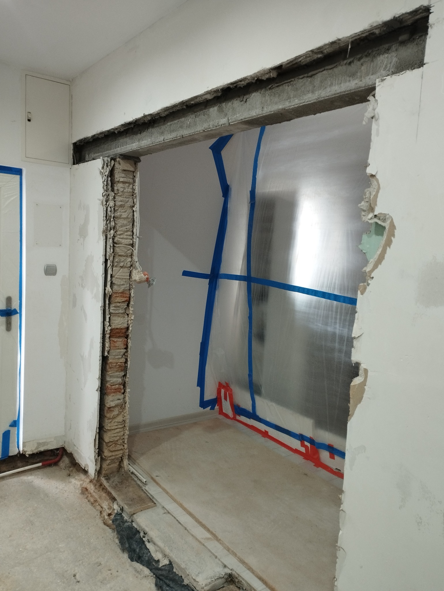 Demontaż ściany działowej z odsłoniętą cegłą, zabezpieczona folią i taśmą malarską, widoczne pozostałości po starej futrynie.