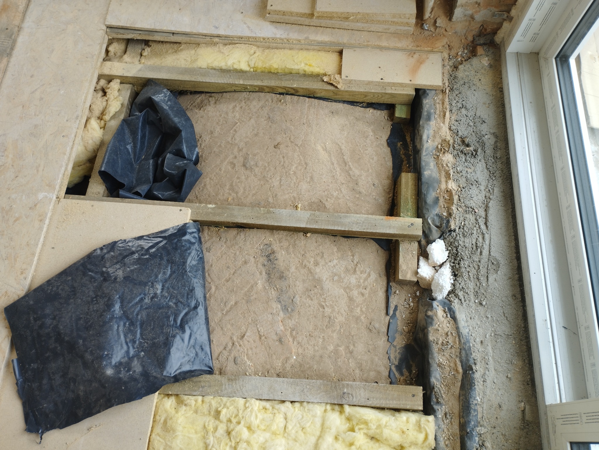 Przygotowanie podłoża pod wylewkę: izolacja termiczna z wełny mineralnej, folia budowlana, drewniane legary i styropian przy oknie.