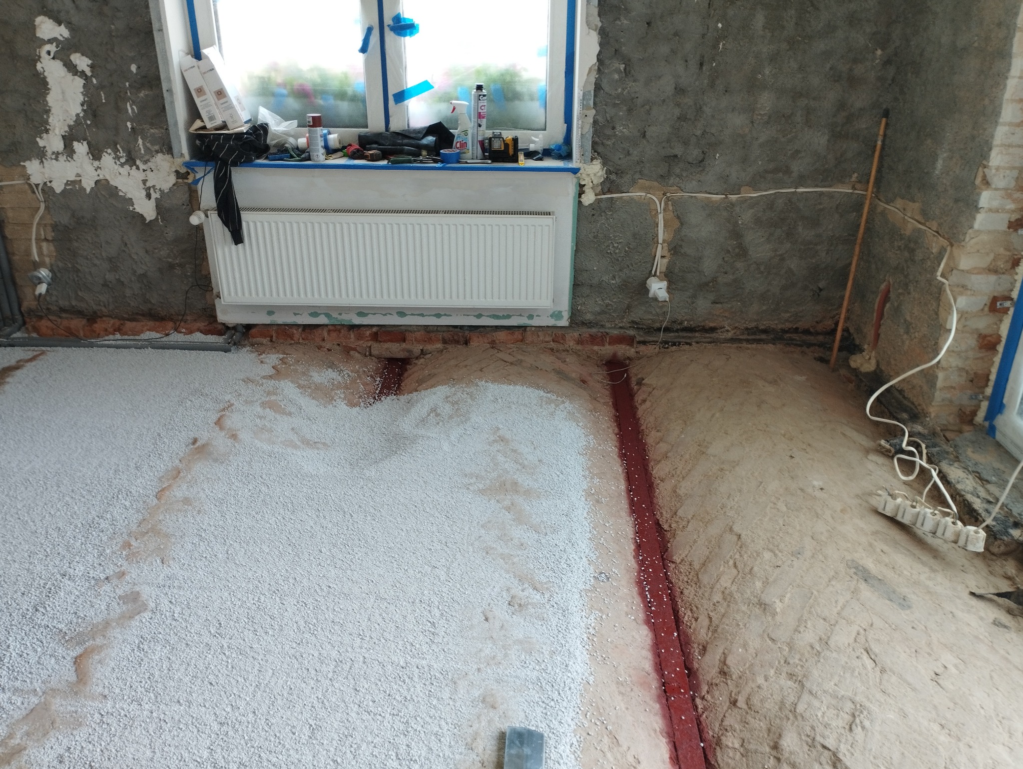 Nierówna podłoga w trakcie wylewania izolacji z białego granulatu, widoczne ślady po instalacjach w podłożu, ściany w surowym stanie, okno zabezpieczone folią malarską, grzejnik zamontowany.