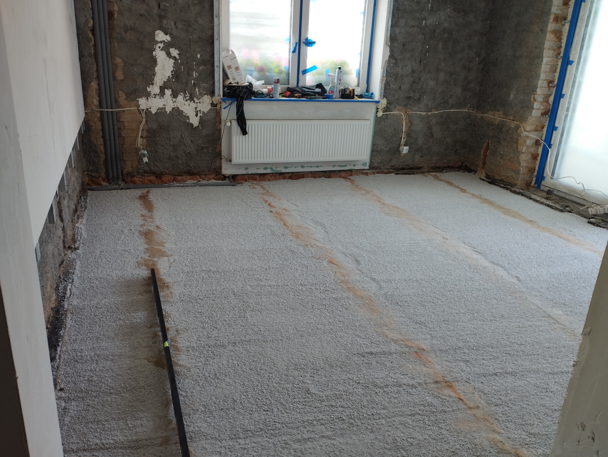 Wyrównany podkład podłogowy z widocznymi śladami po łatach, surowe ściany w tle, okno z osprzętem i grzejnik.