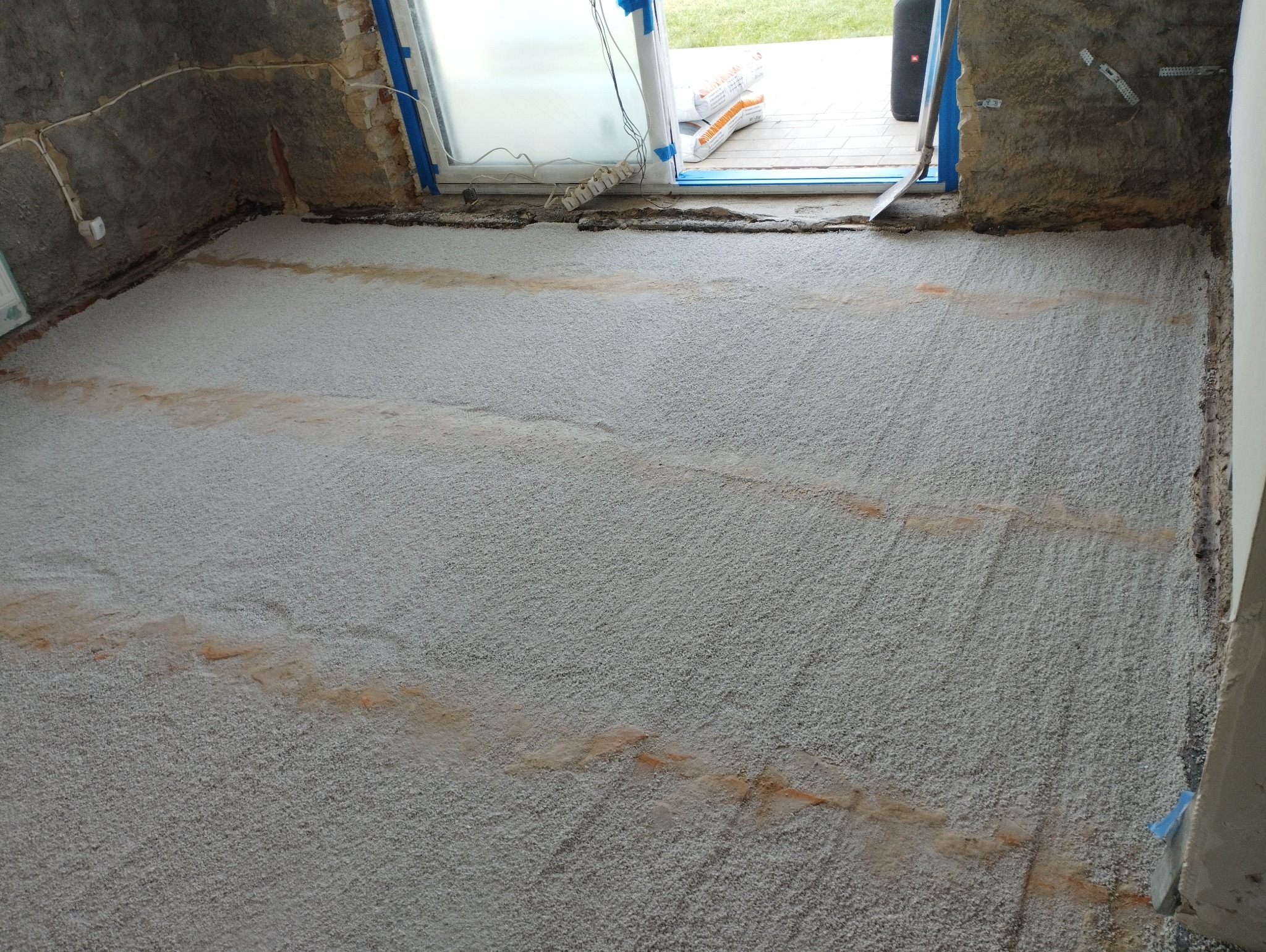 Wyrównana, szara, chropowata powierzchnia podłogi z widocznymi smugami zabrudzeń, przygotowana pod dalsze prace wykończeniowe, przy oknie widoczne worki z materiałem i przedłużacz.