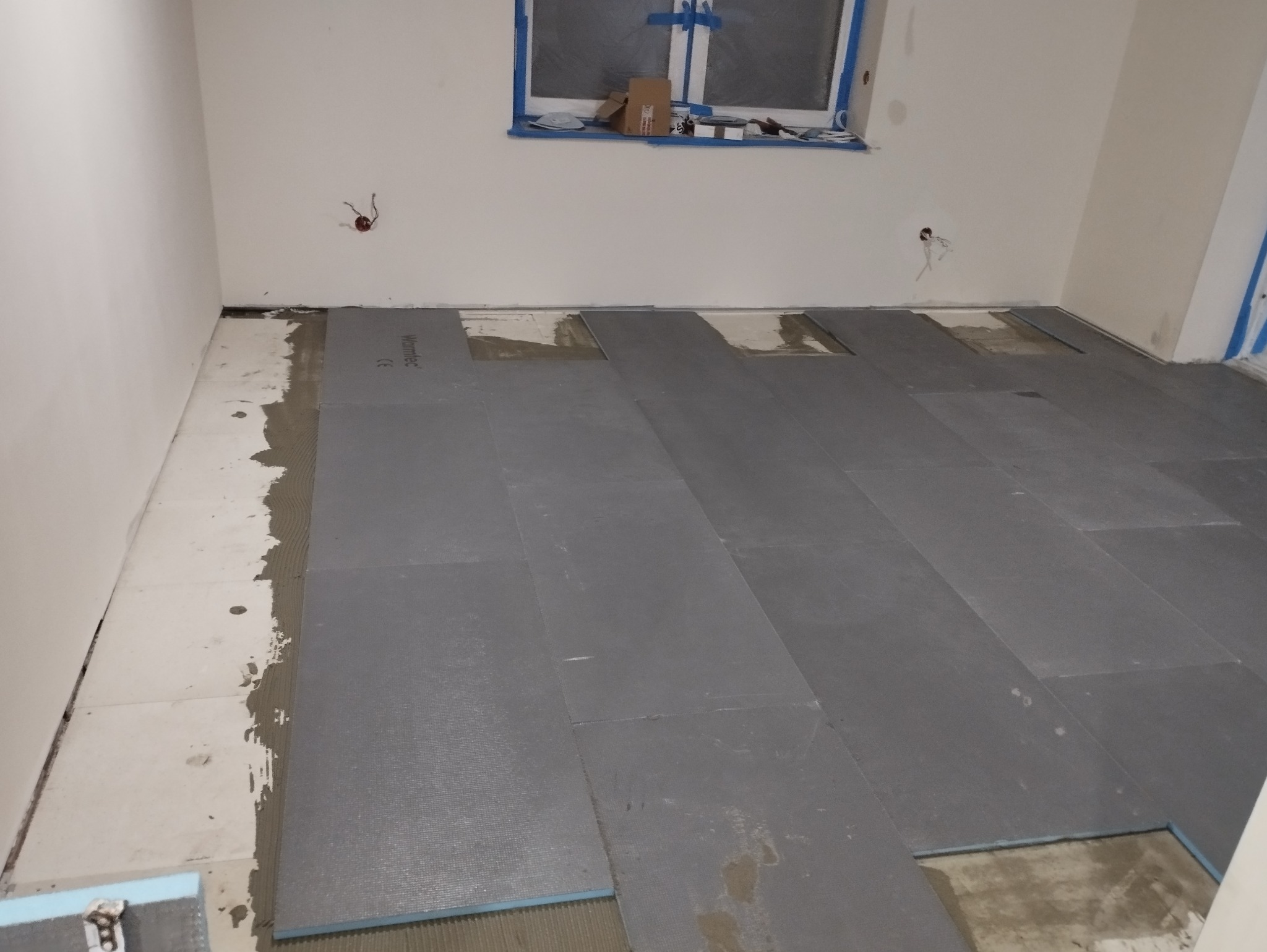 Układanie paneli podłogowych w pomieszczeniu, widoczne płyty izolacyjne Warmtec i warstwa kleju na podłodze, okno zabezpieczone taśmą malarską.