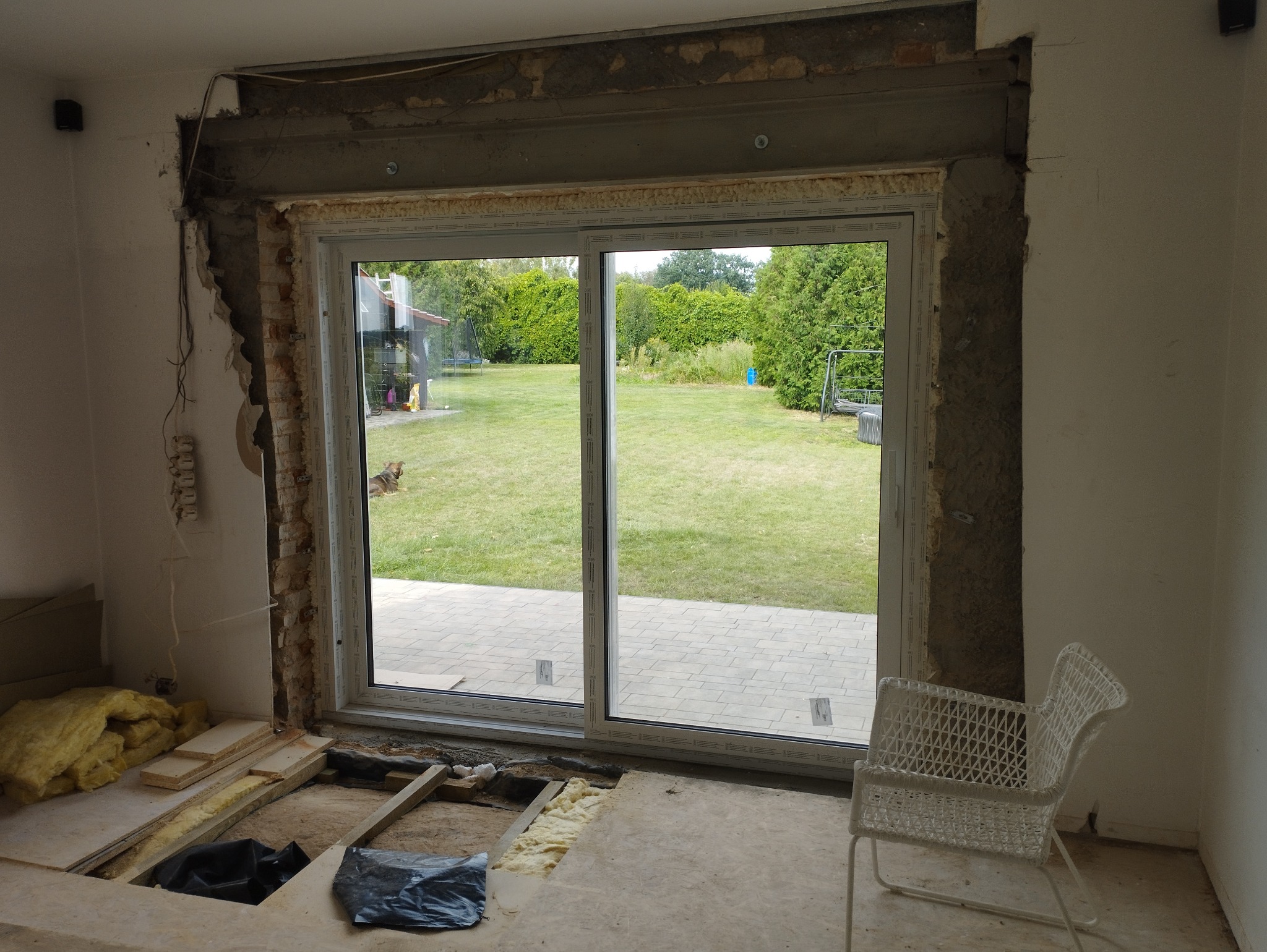 Wnętrze w trakcie remontu: zamontowane okno przesuwne z widokiem na trawnik, odsłonięta cegła i instalacja elektryczna, materiały izolacyjne na podłodze.
