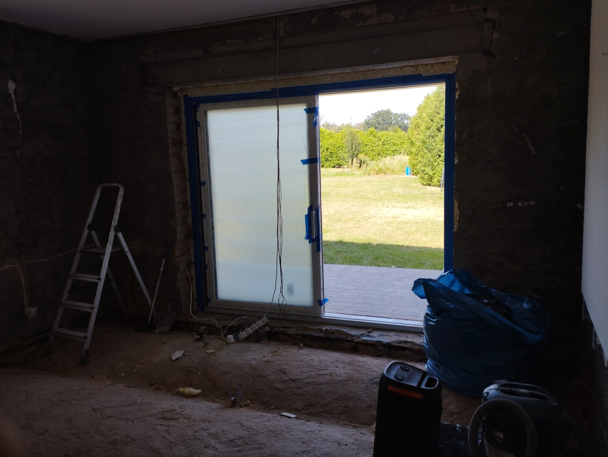 Wnętrze w trakcie remontu z odsłoniętymi ścianami, zabezpieczonymi oknami folią i taśmą malarską, drabina oparta o ścianę, widoczne worki na gruz i odkurzacz.
