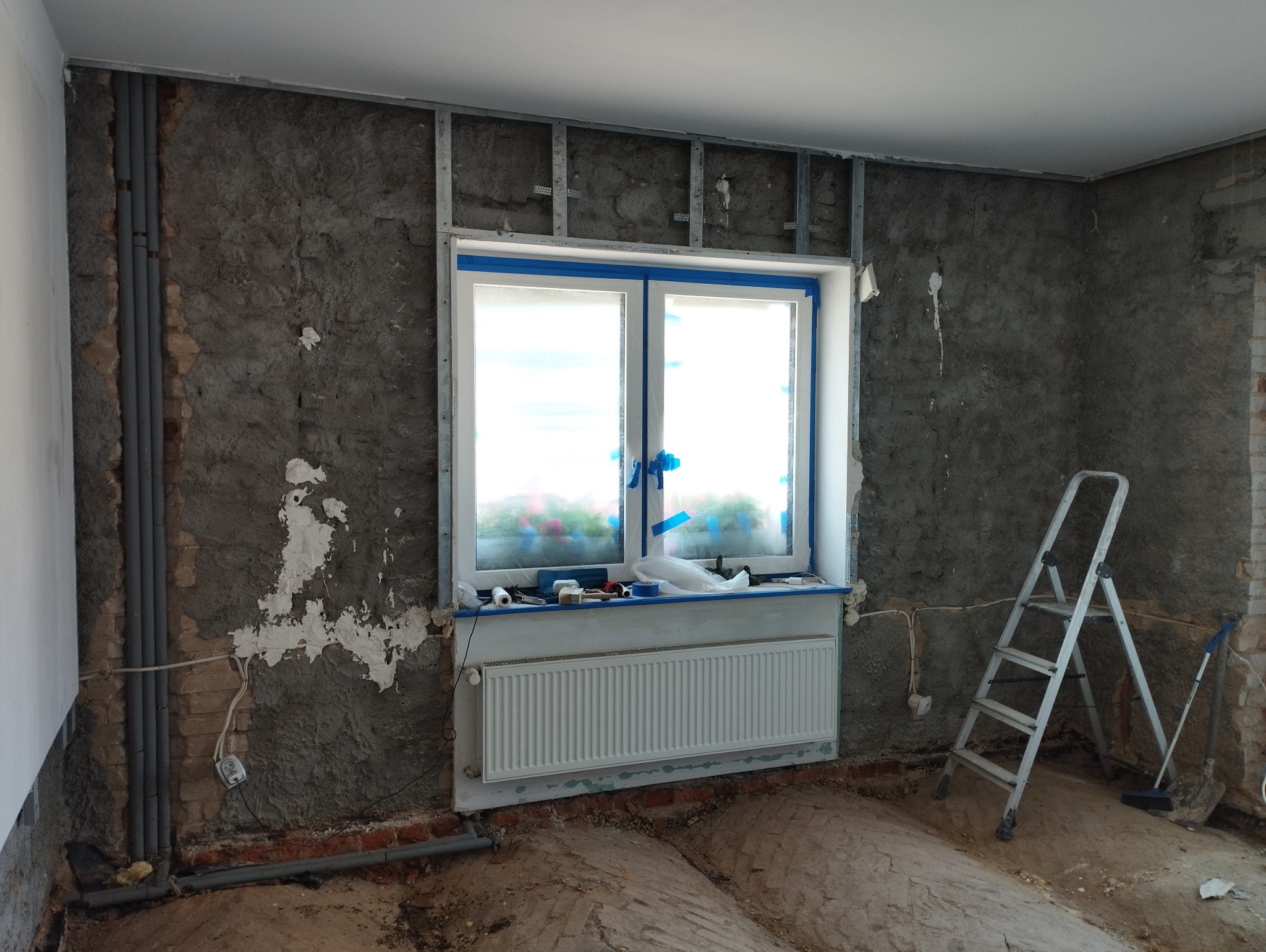 Pokój w trakcie remontu: surowe ściany z widocznymi instalacjami, okno zabezpieczone taśmą malarską, grzejnik, drabina i narzędzia na podłodze.