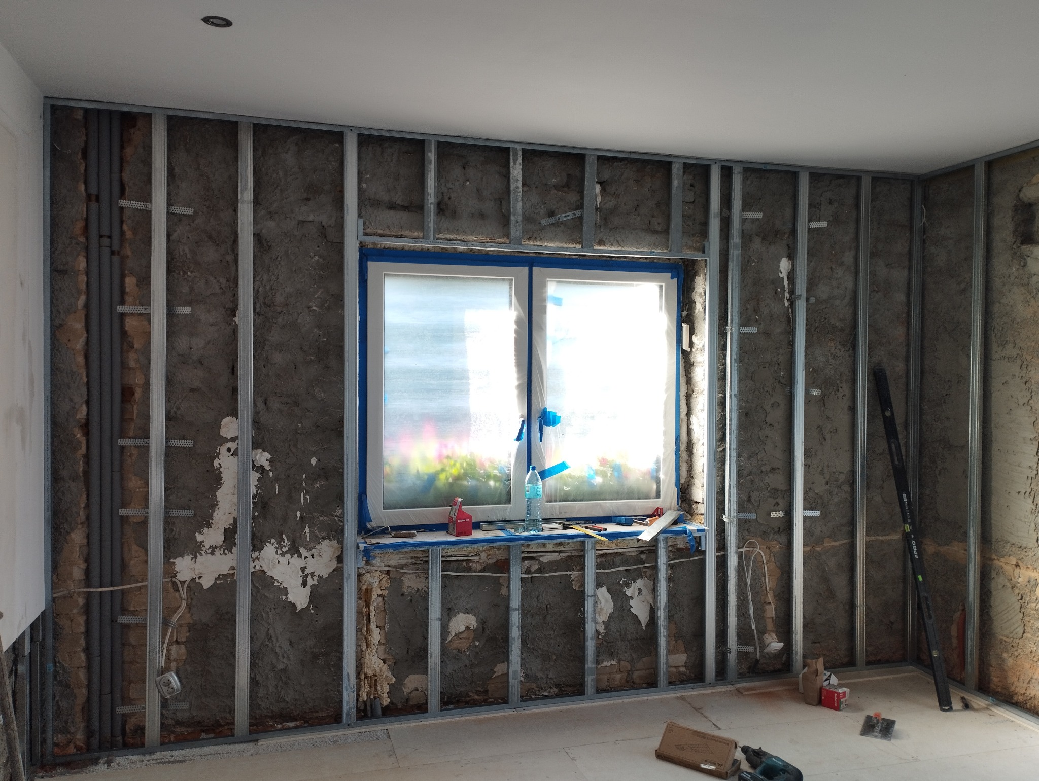 Wnętrze w trakcie remontu: metalowa konstrukcja ścian działowych, okno zabezpieczone folią malarską, widoczne instalacje, poziomica oparta o ścianę, narzędzia na podłodze.