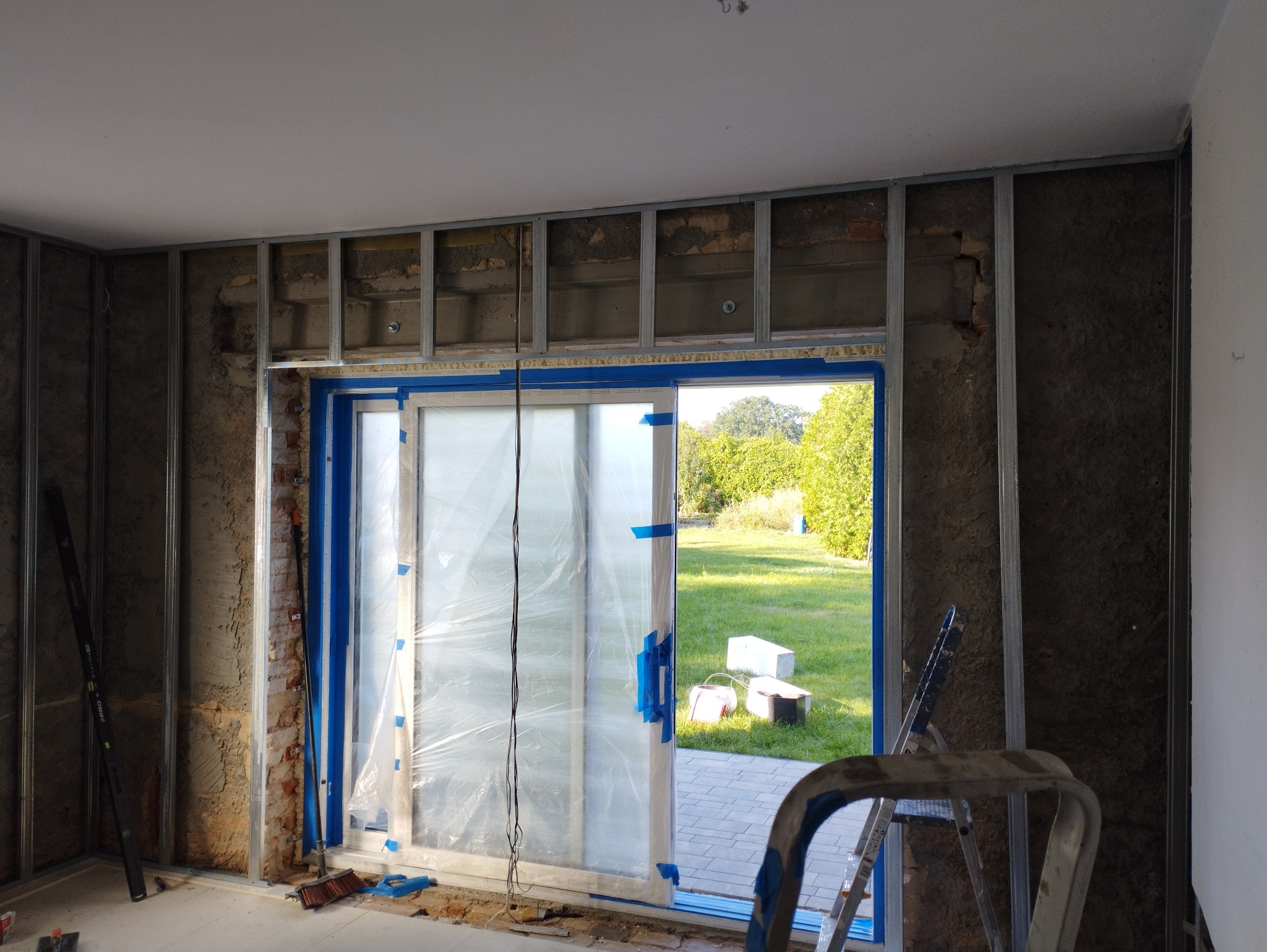 Wnętrze w trakcie remontu z widoczną konstrukcją metalową ścian działowych, zabezpieczonymi folią drzwiami balkonowymi z widokiem na ogród, oraz drabina aluminiowa.