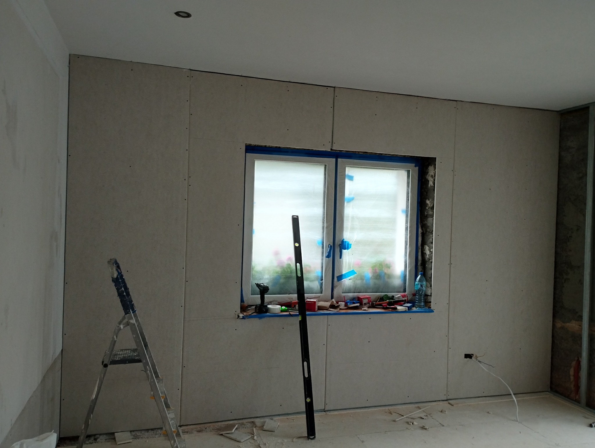 Pokój w trakcie prac wykończeniowych: ściany obłożone płytami gipsowo-kartonowymi, okno zabezpieczone taśmą malarską, poziomica oparta o ścianę, drabina rozstawiona w rogu, widoczne narzędzia...