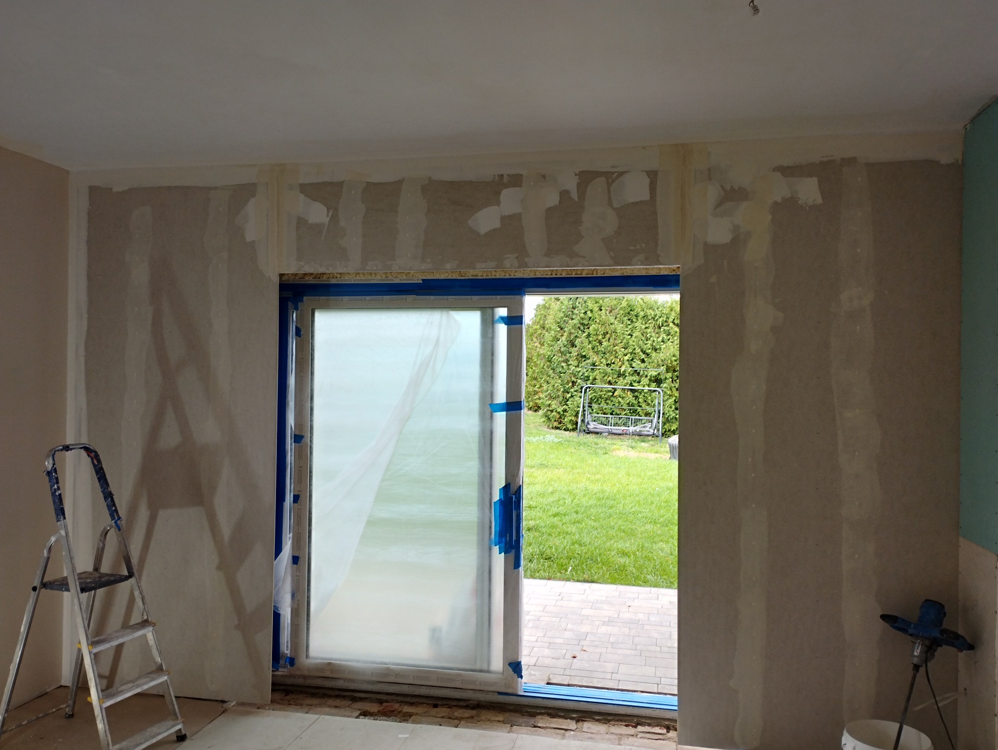 Pokój w trakcie prac wykończeniowych: ściany po nałożeniu warstwy szpachli, okno przesuwne zabezpieczone folią i taśmą, drabina aluminiowa i mieszadło do zaprawy budowlanej