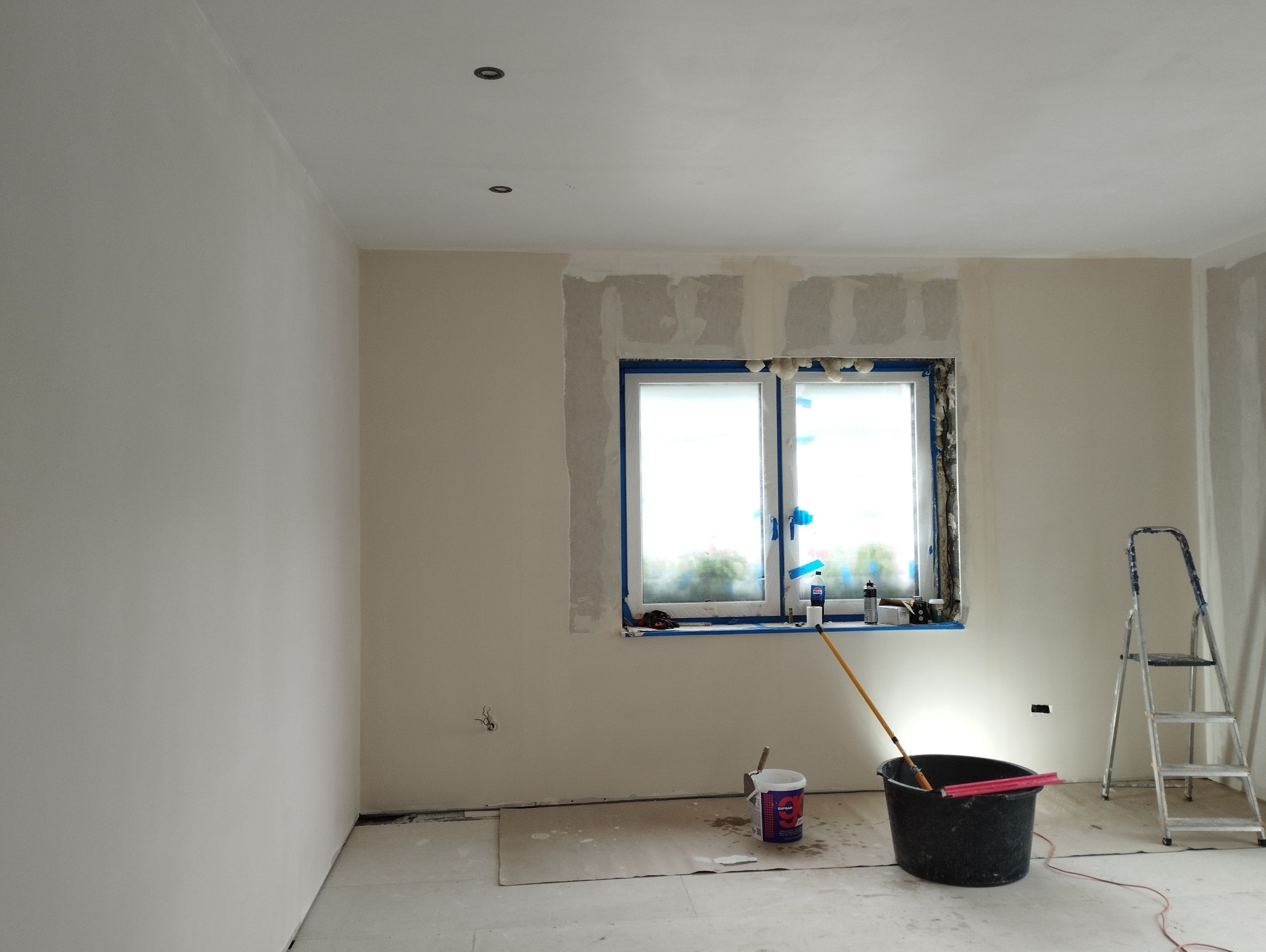 Pokój w trakcie remontu: ściany po szpachlowaniu, zabezpieczone okno folią malarską, wiadro z farbą, drabina aluminiowa i widoczne ślady prac remontowych.