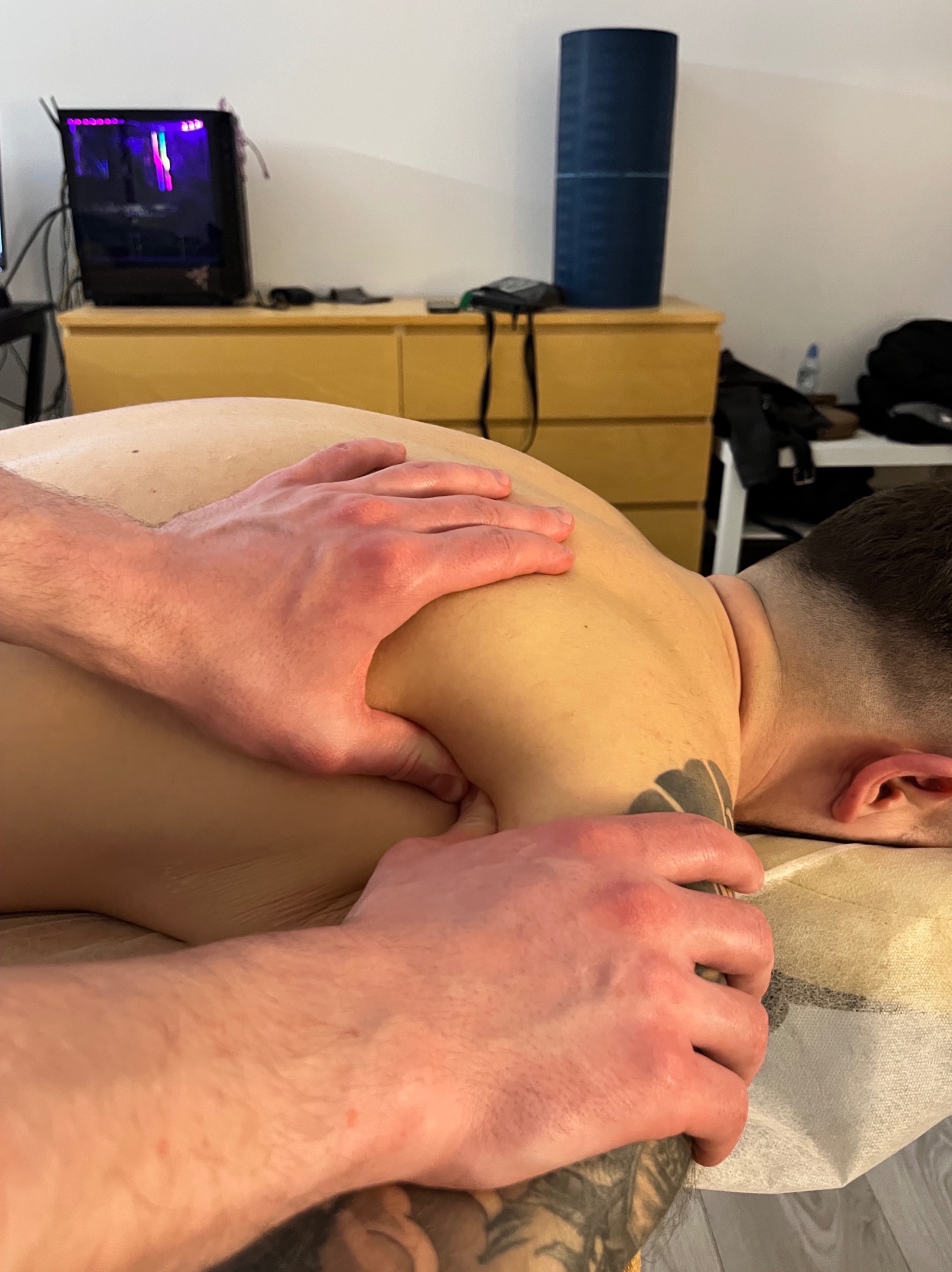 Mężczyzna leży na brzuchu, a terapeuta uciska mu mięśnie górnej części pleców i ramion. Widoczny tatuaż na ramieniu pacjenta i elementy wyposażenia pokoju.