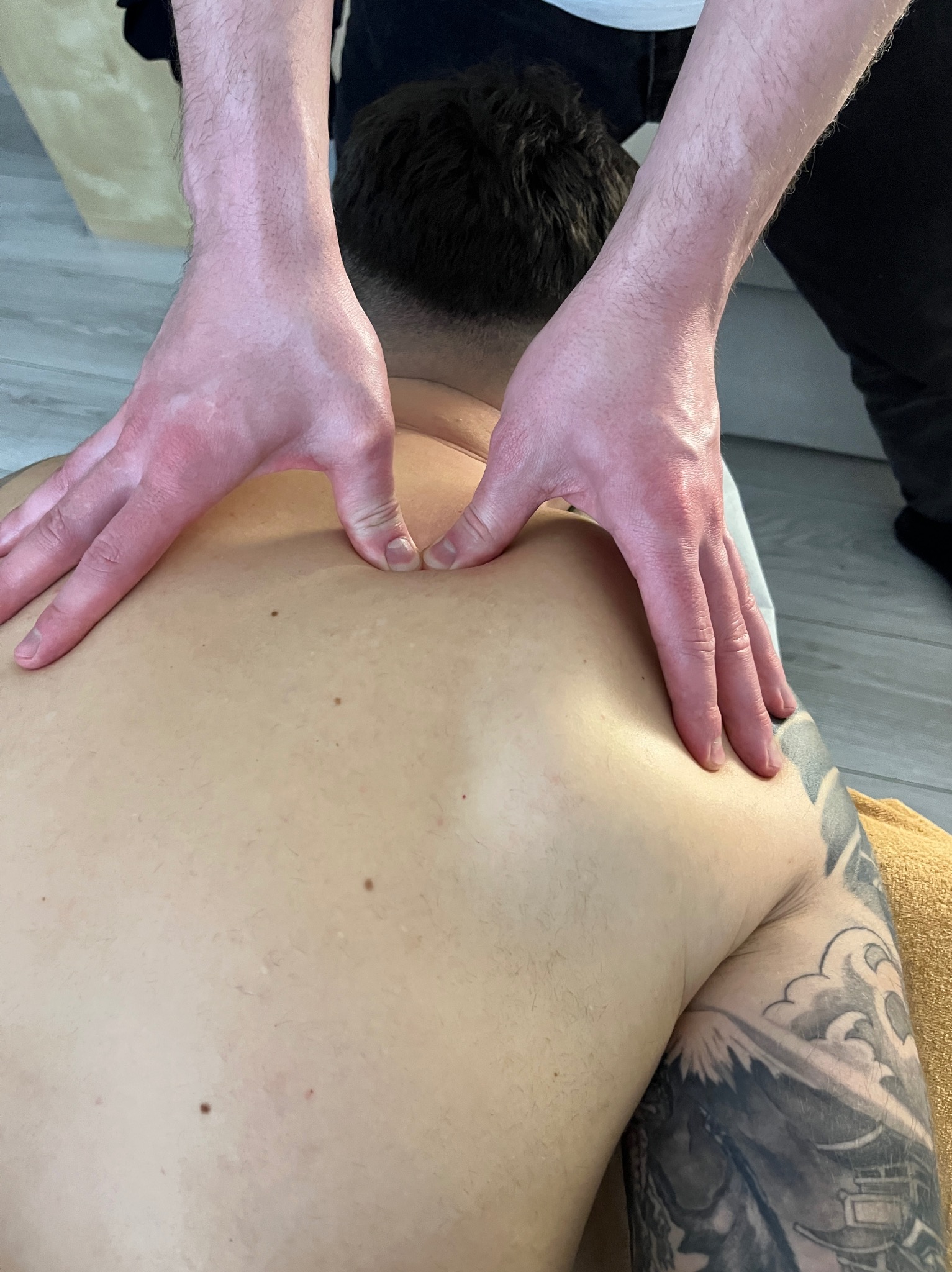 Terapia manualna: Dłonie terapeuty uciskają plecy pacjenta z tatuażem na ramieniu, leżącego na żółtym ręczniku. Ujęcie z góry.