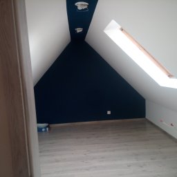 ARKADIUSZ OCH - Poddasze po malowaniu: białe skosy i granatowa ściana szczytowa, drewniana podłoga. Widoczne okno dachowe i wystające kable. Częściowo widoczne drzwi.