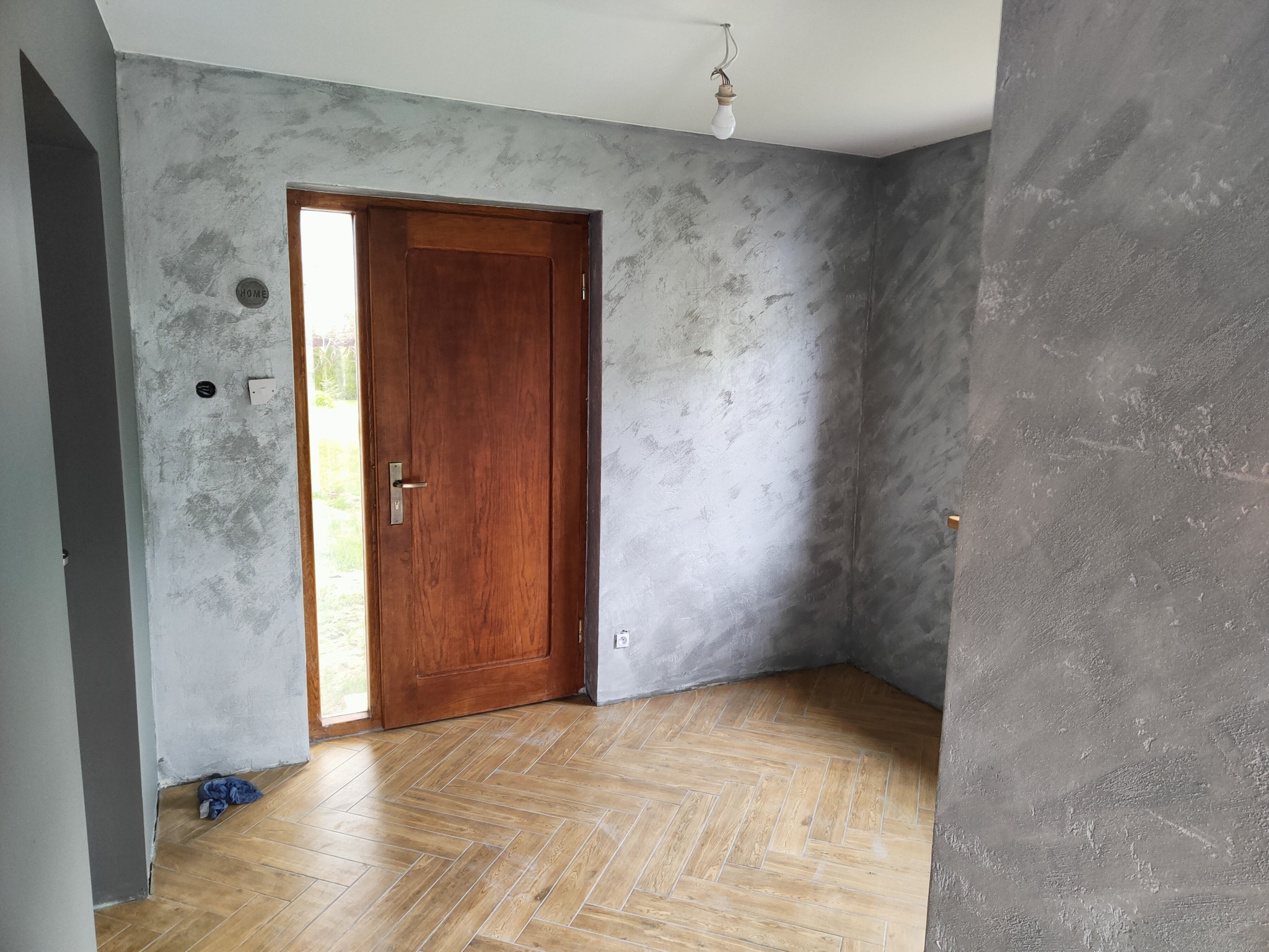 Wnętrze po gładzi dekoracyjnej w odcieniach szarości, z drewnianą podłogą ułożoną w jodełkę i otwartymi drzwiami z brązowego drewna. Widoczna surowa żarówka.