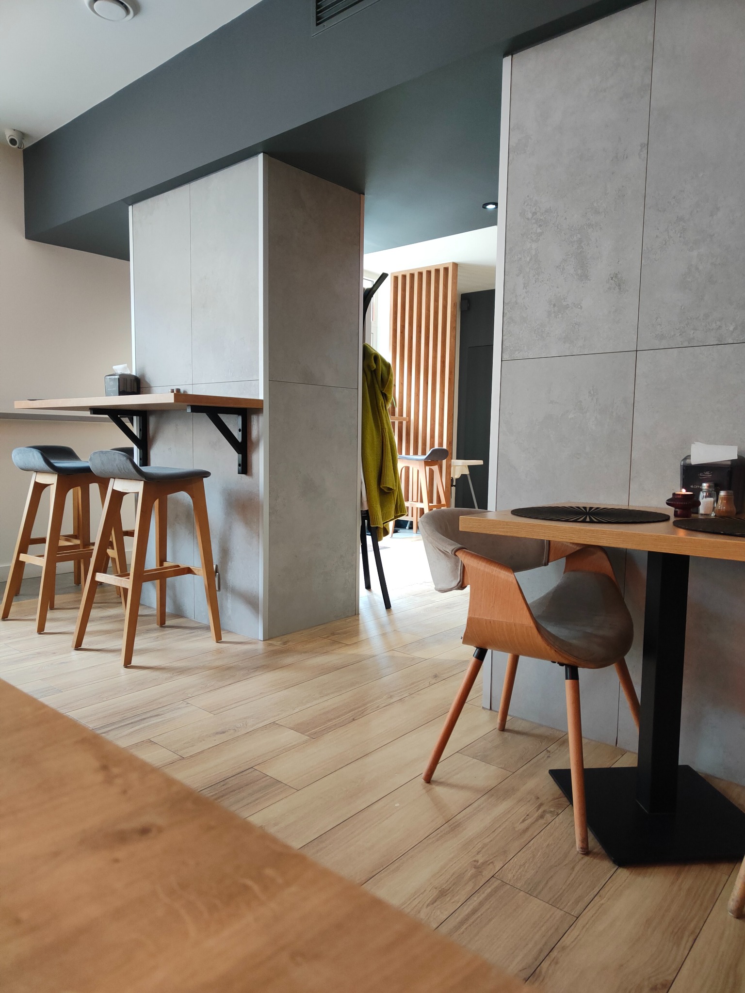 Nowoczesne wykończenie wnętrza kawiarni: drewniane podłogi, szare płytki ścienne, minimalistyczne stołki barowe i stoliki z drewnianymi blatami.