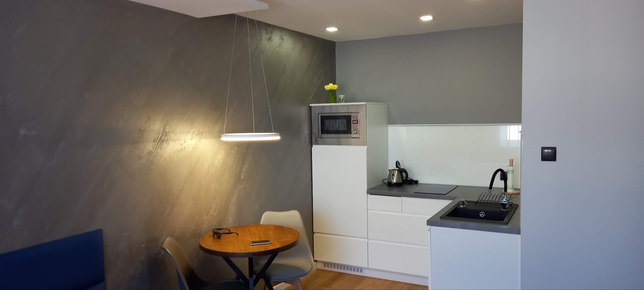 Nowoczesna kuchnia z szarą ścianą o fakturze betonu, okrągłym stołem i minimalistycznym oświetleniem. Białe meble kuchenne i czarny zlew.