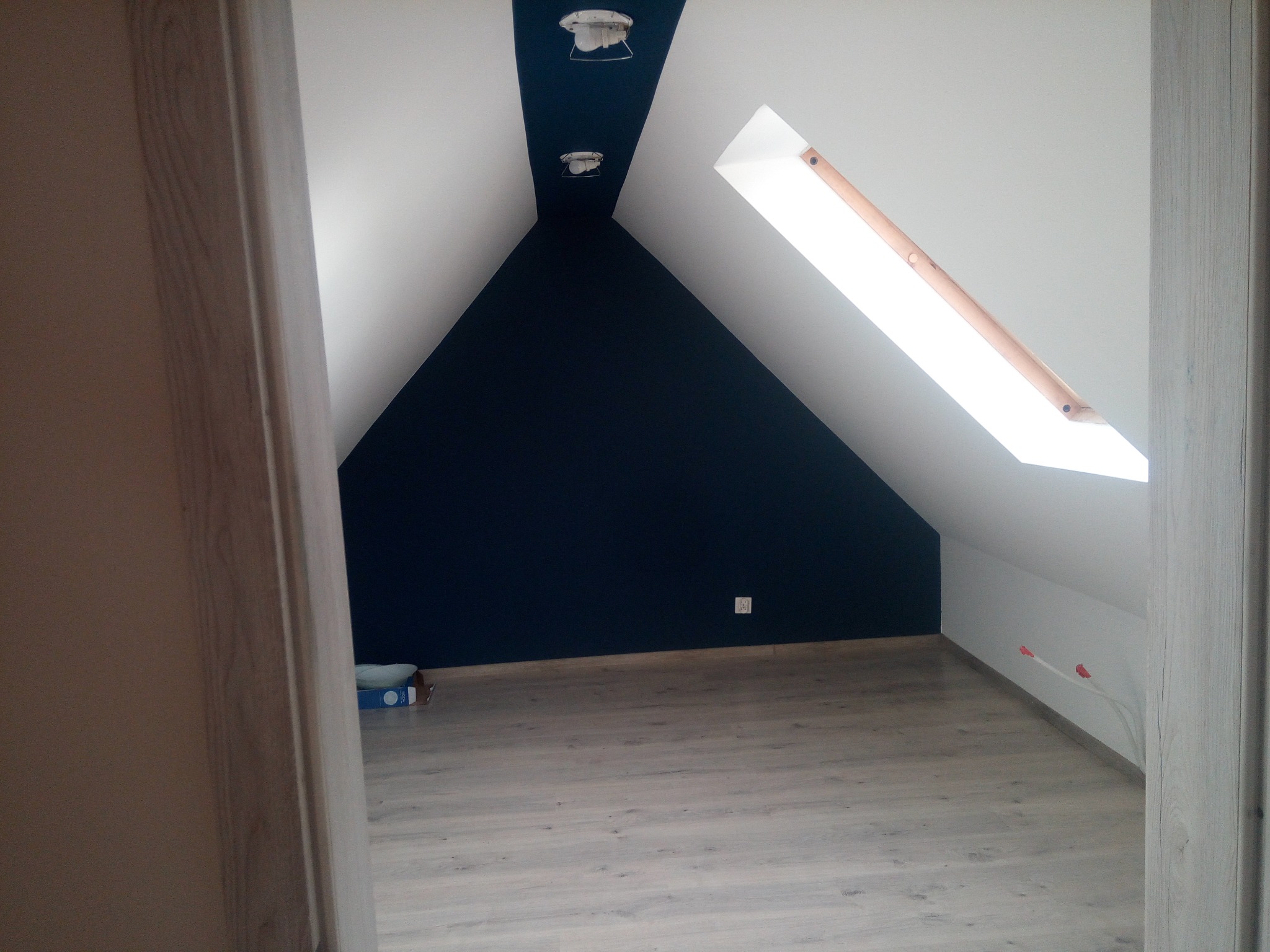Poddasze po malowaniu: białe skosy i granatowa ściana szczytowa, drewniana podłoga. Widoczne okno dachowe i wystające kable. Częściowo widoczne drzwi.