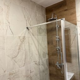 NUD Bud NAZAR HLYNKA - Wykończona kabina prysznicowa z marmurowymi i drewnopodobnymi płytkami w nowoczesnej łazience. Widoczny chromowany zestaw natryskowy i szklane drzwi kabiny.