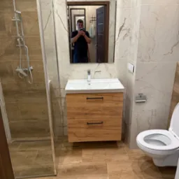 Wykończona łazienka z kabiną prysznicową, umywalką z szafką z drewnopodobnym frontem i toaletą. W lustrze odbija się osoba robiąca zdjęcie.