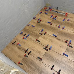 NUD Bud NAZAR HLYNKA - Układanie drewnopodobnych płytek podłogowych w rogu pomieszczenia z użyciem klipsów i klinów poziomujących. Widoczny odpływ liniowy.