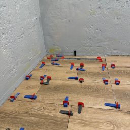 NUD Bud NAZAR HLYNKA - Układanie płytek podłogowych imitujących drewno z użyciem klipsów poziomujących w rogu pomieszczenia z surową, szarą ścianą.