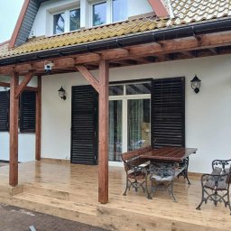 NUD Bud NAZAR HLYNKA - Zadaszony taras z drewnianą podłogą i meblami ogrodowymi. Biała elewacja domu z brązowymi okiennicami i oświetleniem zewnętrznym. Widok na zewnątrz w deszczowy dzień.