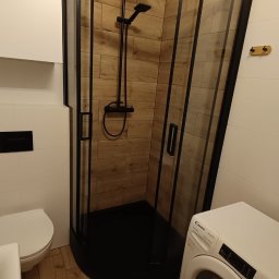 NUD Bud NAZAR HLYNKA - Nowoczesna łazienka po remoncie: kabina prysznicowa z czarną armaturą i drewnopodobnymi płytkami, pralka Candy, biała ceramika. Ujęcie z niskiego kąta.