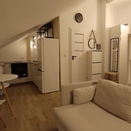 NUD Bud NAZAR HLYNKA - Jasne, minimalistyczne wnętrze poddasza z drewnianą podłogą, białymi meblami i neonowym krzyżem na ścianie. Widoczny aneks kuchenny i część wypoczynkowa.