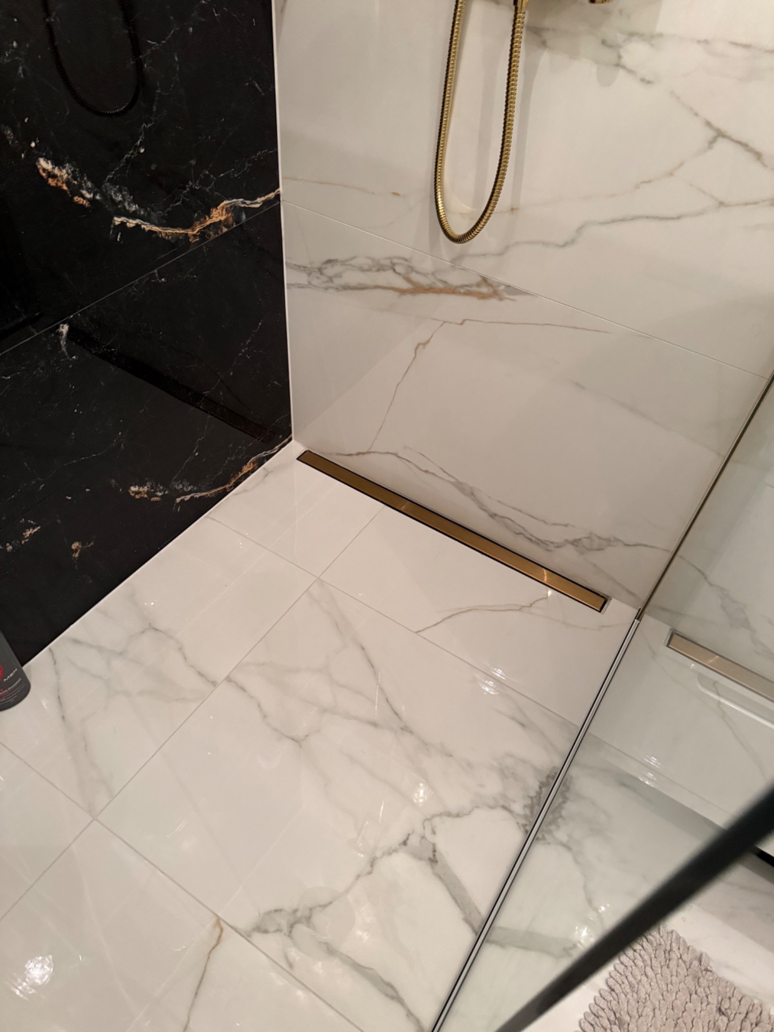 Wykończona kabina prysznicowa z czarno-białymi płytkami imitującymi marmur i złotym odpływem liniowym. Widoczne detale wykończenia i armatura.