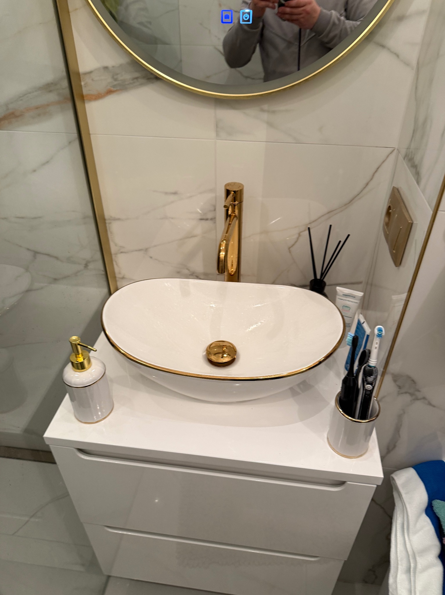 Biała umywalka z armaturą w kolorze złota na szafce w łazience wykończonej marmurowymi płytkami. W tle okrągłe lustro z podświetleniem LED.