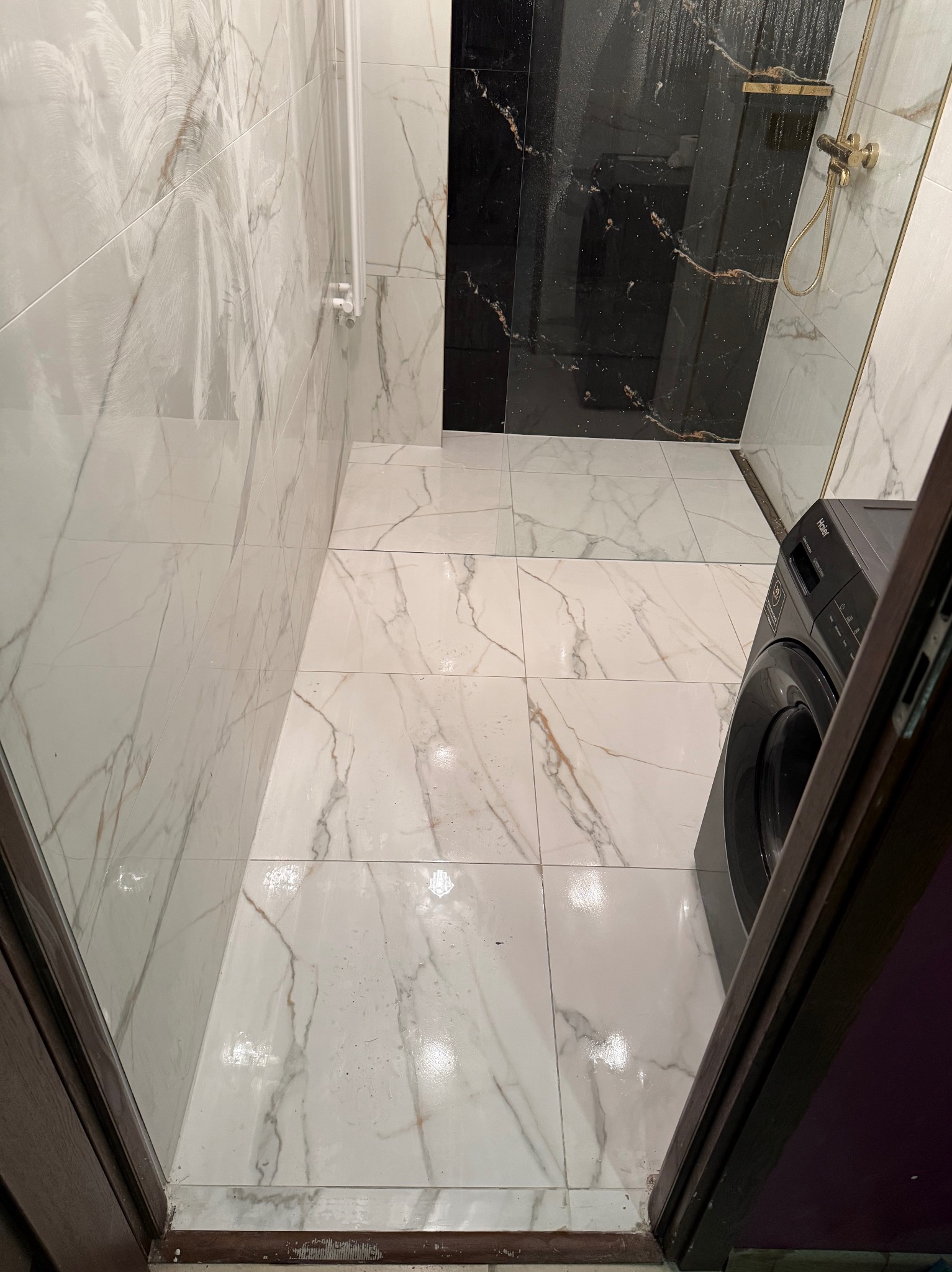 Wykończona łazienka z białymi płytkami imitującymi marmur na ścianach i podłodze, czarną strefą prysznicową i pralką Haier. Widok zza otwartych drzwi.
