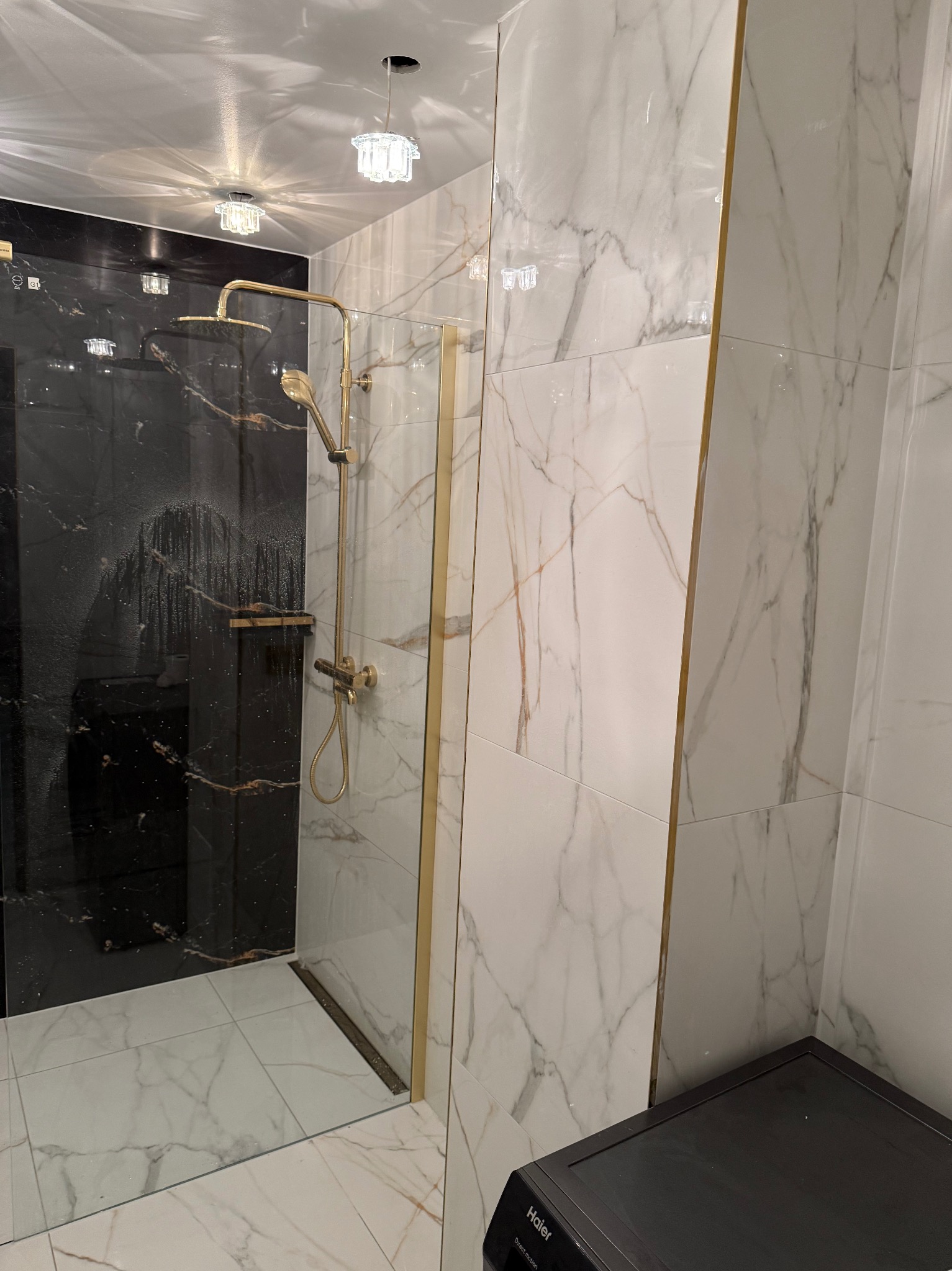Wykończona kabina prysznicowa z czarnymi i białymi marmurowymi płytkami, złotą armaturą i szklanymi drzwiami. Widoczna pralka w rogu.