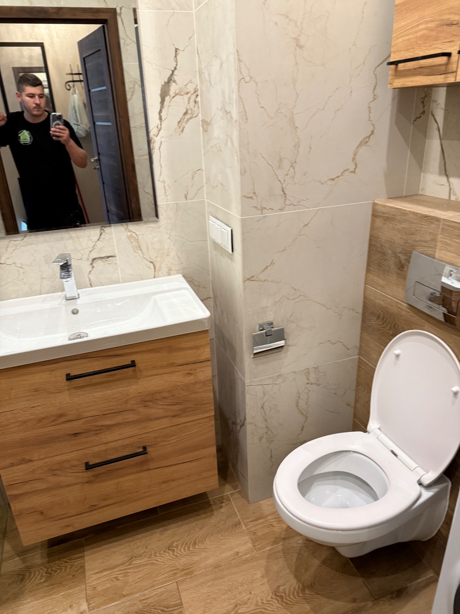 Wykończona łazienka z beżowymi płytkami imitującymi marmur, drewnianą szafką pod umywalką i toaletą. W lustrze odbija się osoba robiąca zdjęcie.